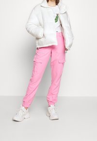 Biała puchowa kurtka, różowe spodnie cargo z elastycznymi mankietami oraz białe, masywne sneakersy. Prosta koszulka z grafiką i zielonym wzorem pod spodem.