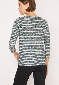 Donna con capelli castani corti che indossa una maglietta a maniche lunghe a righe verde menta, bianca e nera e pantaloni neri, girata di spalle con una mano sul fianco.