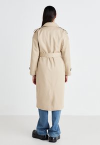 Abrigo trench beige con charreteras, cintura ceñida y abertura trasera, combinado con vaqueros de pierna ancha azules y zuecos negros, visto desde atrás.