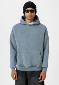 Blauwe hoodie van een katoenmix, met een voorzak, geribbelde boorden en een relaxte pasvorm. Draagt een zwarte beanie en een zonnebril.