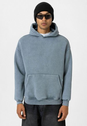 Blauwe hoodie van een katoenmix, met een voorzak, geribbelde boorden en een relaxte pasvorm. Draagt een zwarte beanie en een zonnebril.