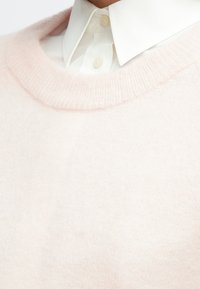 Šviesiai rožinis, minkštas megztinis su paprastu dizainu ir apvalia iškirpte, sluoksniuotas ant balto marškinių su apykakle. Matomas tekstūruotos medžiagos detalė.