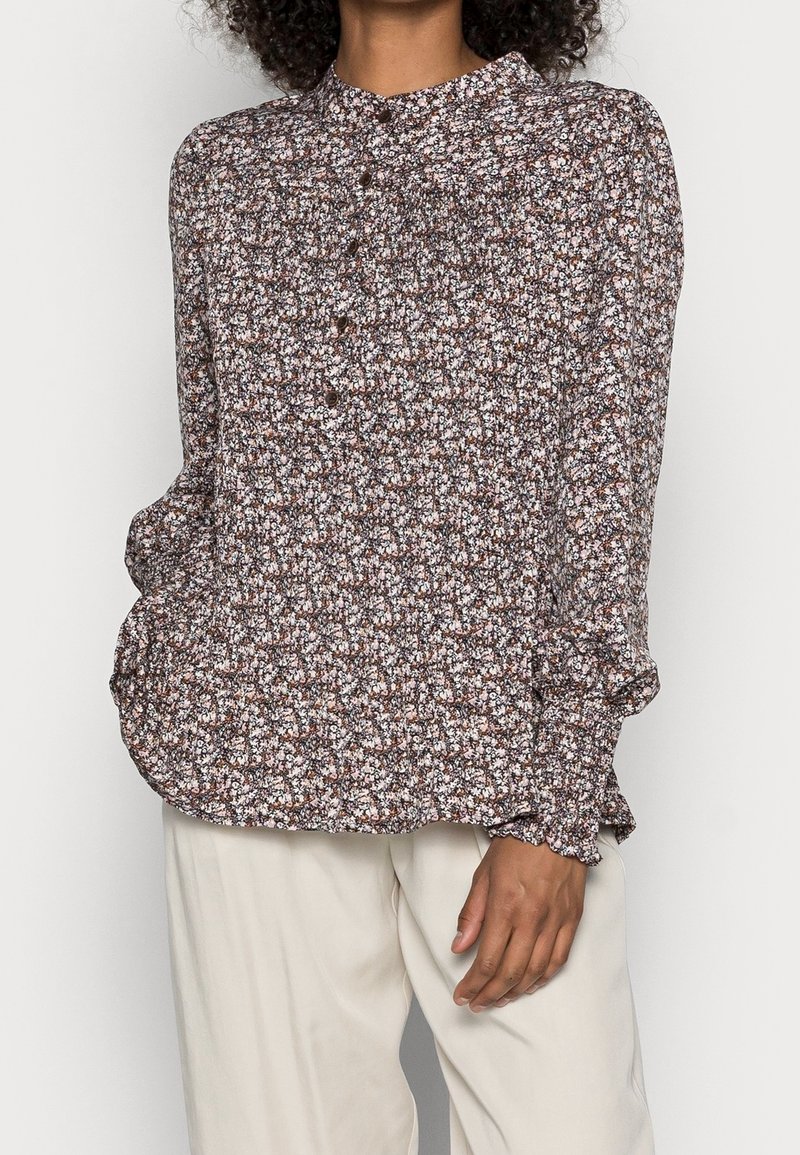 Femme portant un chemisier boutonné à manches longues avec un motif floral marron, blanc et rose, associé à un pantalon beige clair.