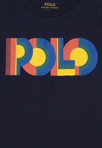 Námorně modrá látka s barevnými geometrickými písmeny tvořícími slovo "POLO" v žluté, modré, zelené a červené barvě, s textem "POLO RALPH LAUREN" nad ním.