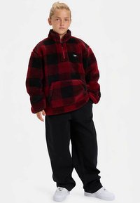 Pulôver de fleece aos quadrados vermelhos e pretos com gola e bolso frontal com fecho de correr, combinado com calças largas pretas e ténis brancos.