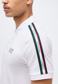 Άνδρας που φοράει λευκό polo πουκάμισο EA7 Emporio Armani με χαρακτηριστικές μαύρες, πράσινες, λευκές και κόκκινες ρίγες στον ώμο και το λογότυπο στο στήθος.