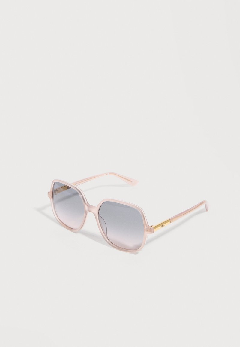 Lunettes de soleil à monture carrée translucide rose avec des verres à dégradé gris et des accents dorés sur les branches, posées sur une surface blanche.