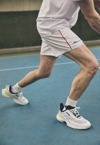 Sport shoes with a white mesh upper, navy accents, and a neon green detail on the sole. Worn with white athletic shorts and socks.