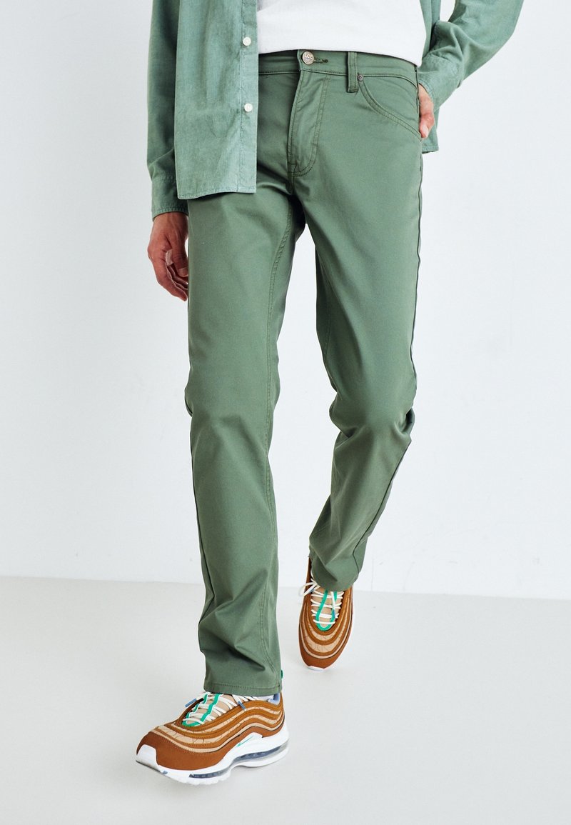 Pantalons vert olive en mélange de coton, avec une coupe droite, cinq poches et des détails de couture subtils, associés à des baskets marron.