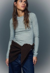 Siva dolgo rokavna majica z oprijeto zasnovo, kombinirana s temno rjavim pletenim kosom, ovitim okoli pasu in zavezanim, nošena preko modrih jeansa.