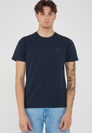 Jonge man met krullend haar, draagt een marineblauw T-shirt en lichtblauwe spijkerbroek, toont tatoeages op zijn linker onderarm tegen een effen achtergrond.