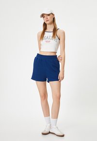 Witte crop top met tekst, blauwe shorts en witte sneakers. Model draagt een witte pet en witte sokken, staande tegen een effen achtergrond.