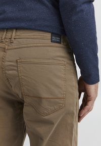 Braune Baumwollhosen mit einem geraden Design, ausgestattet mit einer Gesäßtasche mit spitzem Deckel und einem schwarzen Etikett, das "FQ/1924 ORIGINAL" trägt.