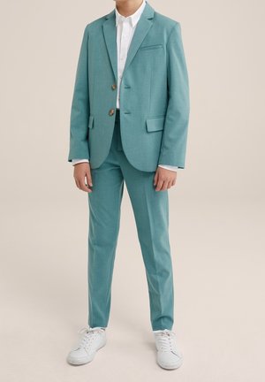 Garçon portant un costume bleu sarcelle à deux boutons avec une chemise blanche et des baskets blanches, debout devant un fond beige uni.