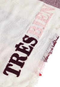 Couverture tissée avec des bords fringés, arborant le texte brodé "TRÈS BIEN" en bourgogne foncé et en rose clair sur un fond blanc.