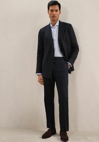 Sacou bleumarin cu două rânduri de nasturi și rever la fante, purtat cu o cămașă cu dungi albastru deschis și pantaloni asortați. Pantofii maro completează ținuta.