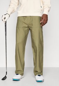 Pantaloni de golf khaki cu design drept, realizați dintr-un material din amestec de bumbac. Dispune de buzunare laterale și sunt asortați cu pantofi de golf albi.
