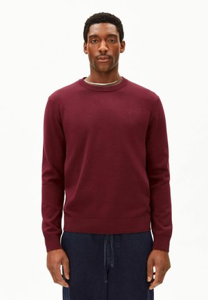 Homme portant un pull bordeaux uni à manches longues et un pantalon bleu foncé, debout devant un fond blanc uni.