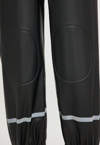 Svarta vattentäta byxor med vadderade knäpatchar och reflexer i horisontella ränder vid anklarna på vit bakgrund.
