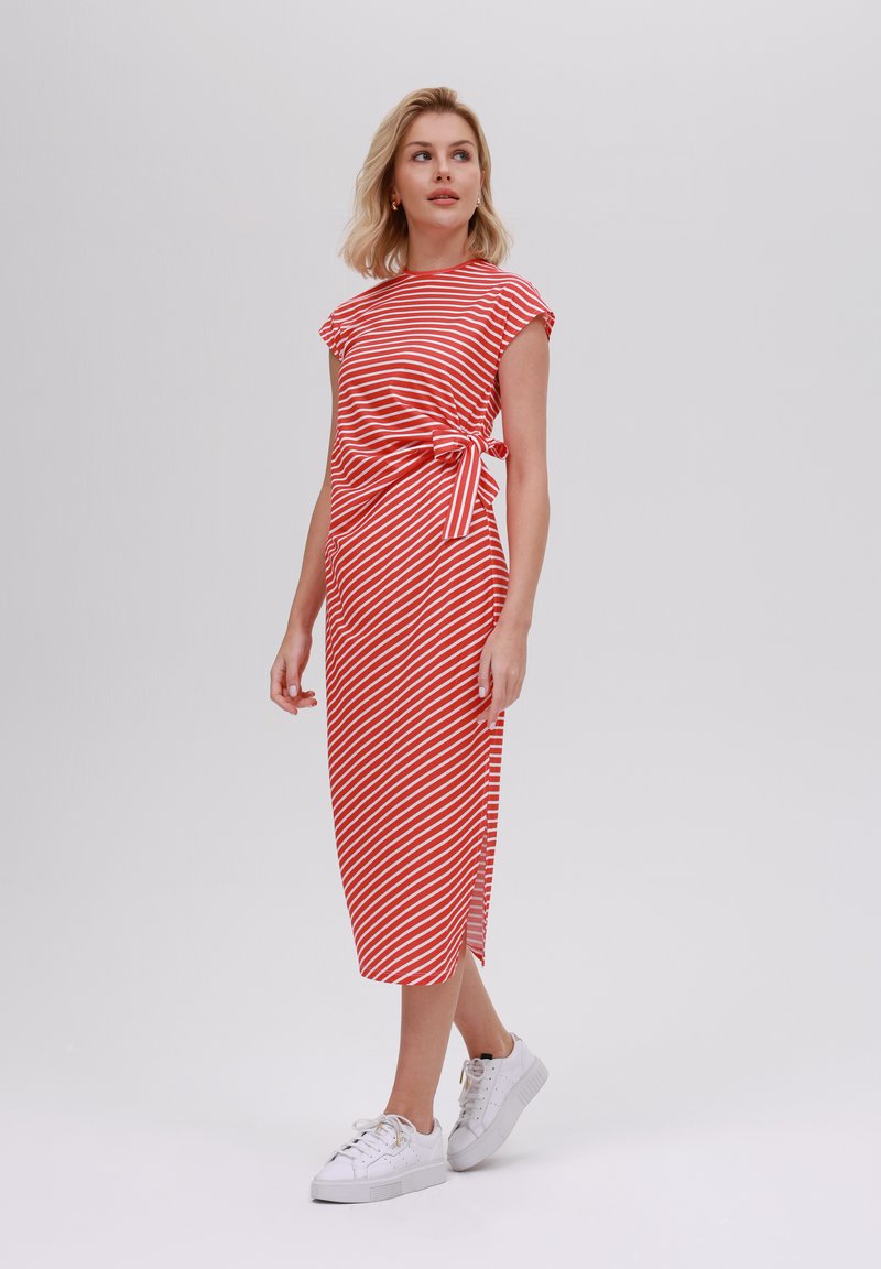 Robe rayée rouge et blanche avec une taille cintrée et un détail de nœud sur le côté, dotée d'une longueur midi et de manches courtes, associée à des baskets blanches.