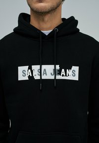 Sudadera negra con capucha y bolsillo frontal, que presenta un gráfico rectangular blanco con la inscripción "SALSA JEANS" en una tipografía gruesa con efecto desgastado.