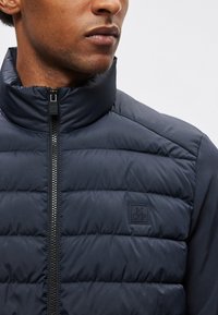 Mørkeblå dunvest med høj krave, lynlåslukning og quiltet design. Har et lille logo-patch nær lynlåsen på brystet.