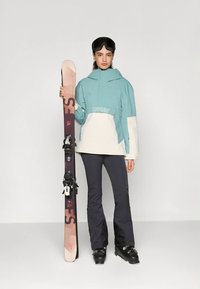 Skidrakt med en lyseblå og hvit vanntett jakke, svarte skibukser, svarte skistøvler og et brunt skipar med bindinger.