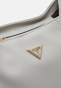 Harmaa teksturoitu nahkakäsilaukku, jossa on kultainen kolmion muotoinen logo-plaakki. Vetoketjusulku, jossa näkyvä sauma reunoilla.