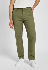 Oliwkowe bawełniane spodnie o prostej nogawce, z przednią kieszenią, metalowym zapięciem na guzik, noszone z białymi trampkami.