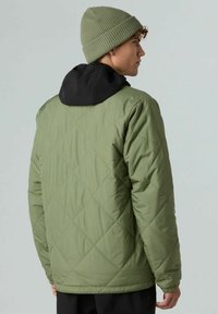 Giovane uomo che indossa una giacca imbottita verde con cappuccio nero e un berretto lavorato a maglia verde oliva, girato di lato su uno sfondo semplice.