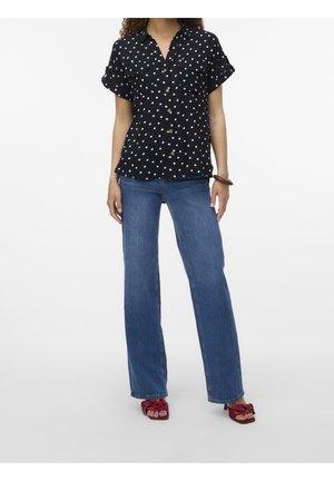 Kvinde iført sort kortærmet bluse med polka-prikker, blå jeans med lige ben, røde sandaler med sløjfer og et brunt armbånd på venstre håndled.