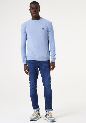 Sudadera de manga larga azul claro con cuello redondo y pequeño logotipo negro, combinada con jeans de corte slim fit azul oscuro y zapatillas con acentos grises.