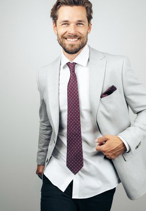 Smiling man in light gray suit jacket, white shirt, maroon patterned tie, and matching pocket square, standing against plain light background.