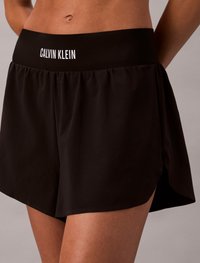 Mustad sportlikud lühikesed püksid laia vööga, millel on valge "CALVIN KLEIN" bränding. Kujunduses on külgmised lõhenemised liikuvuse ja mugavuse tagamiseks.