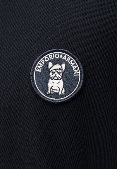 Στρογγυλό κεντημένο πατς σε μαύρο ύφασμα με έναν γαλλικό μπουλντόγκ που φορά μπερέ, περιτριγυρισμένο από το κείμενο «EMPORIO ARMANI».