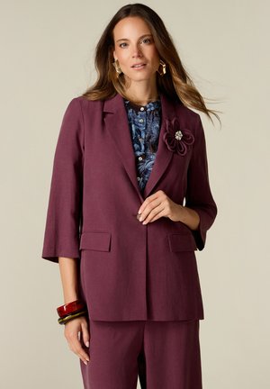 Femme portant un blazer violet avec une broche fleurie, un pantalon assorti, un chemisier bleu à motifs et des bracelets épais à un poignet.