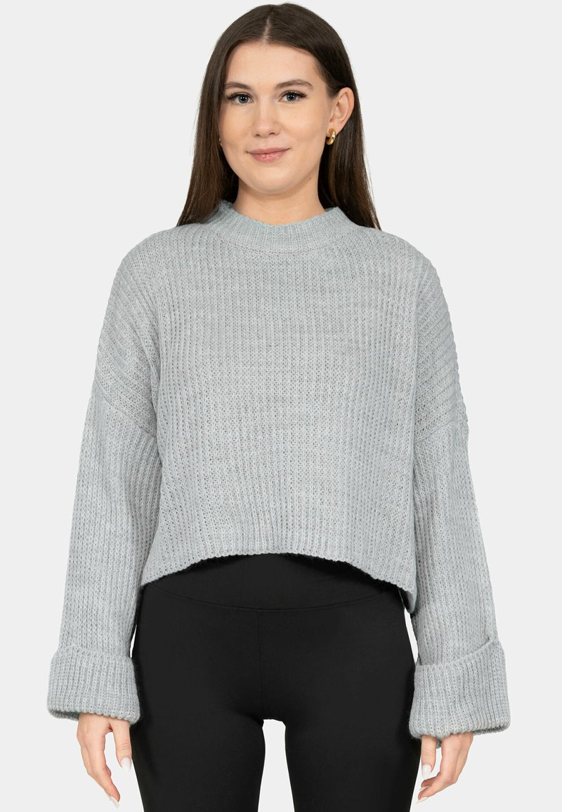 Giovane donna che indossa un maglione grigio chiaro lavorato a maglia, morbido con polsini arrotolati, e leggings neri, in piedi davanti a uno sfondo chiaro uniforme.