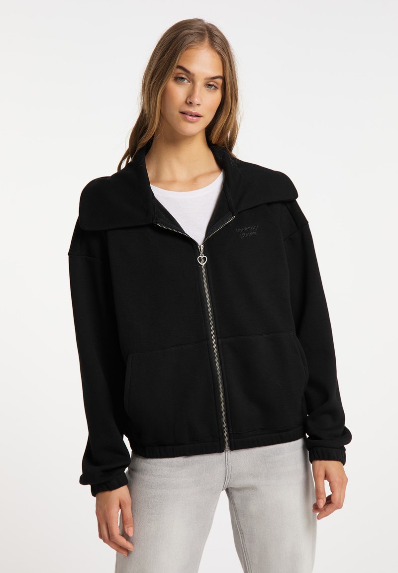 Felpa nera con zip e colletto ampio, realizzata in tessuto morbido, con due tasche frontali e polsini elasticizzati. Indossata sopra una t-shirt bianca.