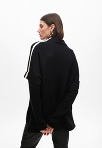 Černý oversized pletený svetr s ohrnutými rukávy a bílými pruhy na ramenou; hladká textura; nošený přes černé kožené kalhoty.