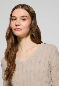 Beige stickad tröja med V-ringning, med kabelmönster och ribbat avslut. Diskret broderad logotyp på bröstet. Mjuk textur.