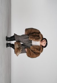 Sieviete valkā brūnu kažokādu jaku, pelēkas platās bikses, melnus zābakus un tur rokās brūnu kažokādas mufu uz vienkārša pelēka fona.