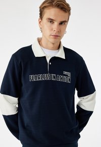 Sudadera navy con cuello y mangas en crema, con texto frontal "SIN MIEDO EN ACCIÓN" y un logotipo. Confeccionada en una suave tela texturizada.