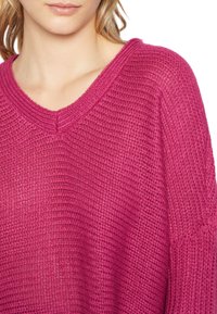 V-ringad tröja i djup rosa med texturerad stickad mönster och sänkta axlar, med ribbade detaljer vid hals och ärmslut.