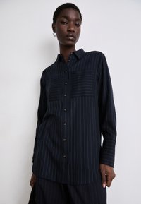 Navy blue button-up shirt with vertical white stripes, two chest pockets, and a collared neckline. Smooth texture, long sleeves.