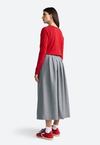 Vrouw in een rood shirt met lange mouwen, een grijze geplooide midi-rok, witte gerimpelde sokken en rode sneakers staat links gericht op een witte achtergrond.