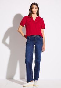 Top rosso a V con maniche corte, abbinato a jeans blu a gamba dritta e sneakers bianche, su uno sfondo neutro.