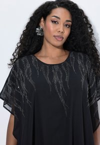 Femme aux longs cheveux bouclés portant de grandes boucles d'oreilles en forme de fleur et une robe noire ornée de motifs de perles scintillantes sur les épaules.