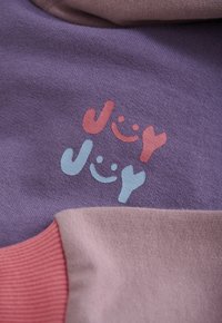 Lila und rosafarbener Stoff mit aufgedrucktem Text "Joy Joy" in Rot und Hellblau, wobei jedes Wort von einem Smiley aus den Buchstaben begleitet wird.