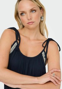 Robe marine avec des liens d'épaule tressés et un motif de feuilles brodé. La texture plissée ajoute de la dimension. Les bras du mannequin sont croisés, des boucles d'oreilles en argent sont visibles.