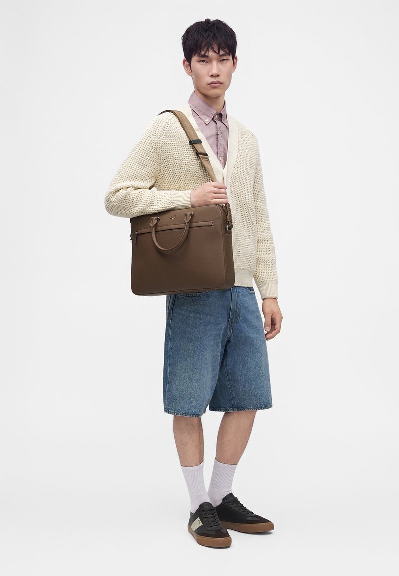 Jeune homme portant un cardigan crème, un short en denim, des baskets noires et portant un sac à bandoulière en cuir marron sur un fond uni.